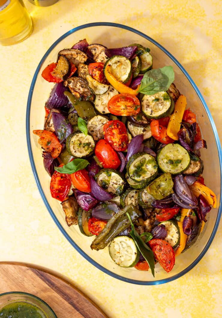 A large oval dish with roasted vegetables;tomatoes, peppers, red onions and courgettes on a pale yellow background.