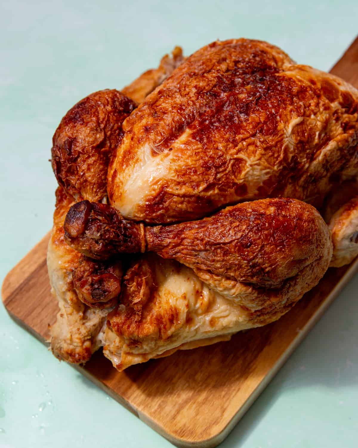 Large golden browned whole chicken on a wooden board on a pale green background.