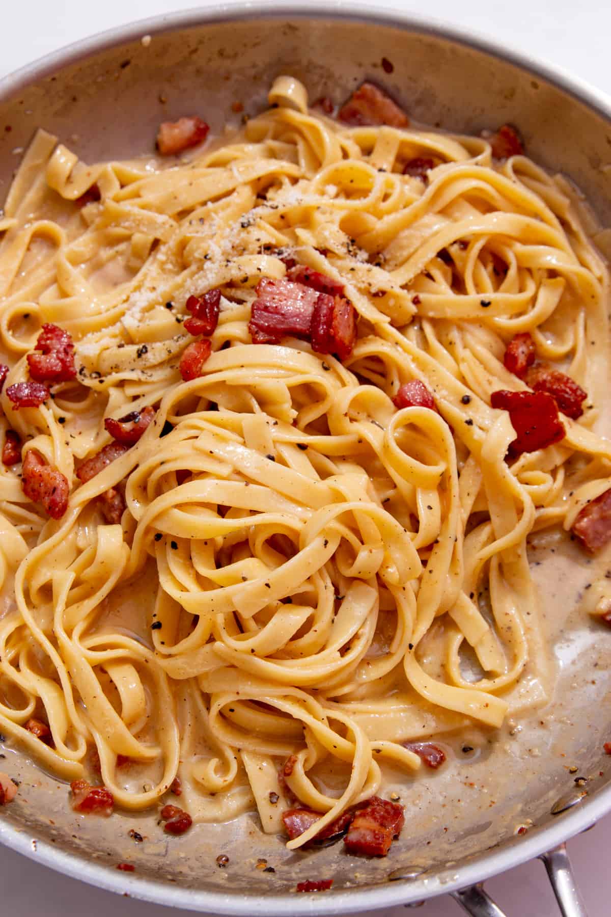 Tagliatelle in a large stainless steel pan with pancetta and parmesan topping.