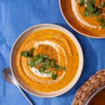 2 bowls with carrot soup garnished with a swirl of cream and coriander on a blue background.