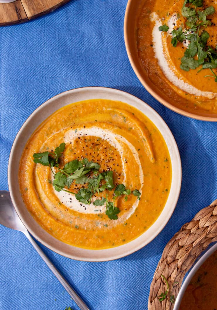 2 bowls with carrot soup garnished with a swirl of cream and coriander on a blue background.