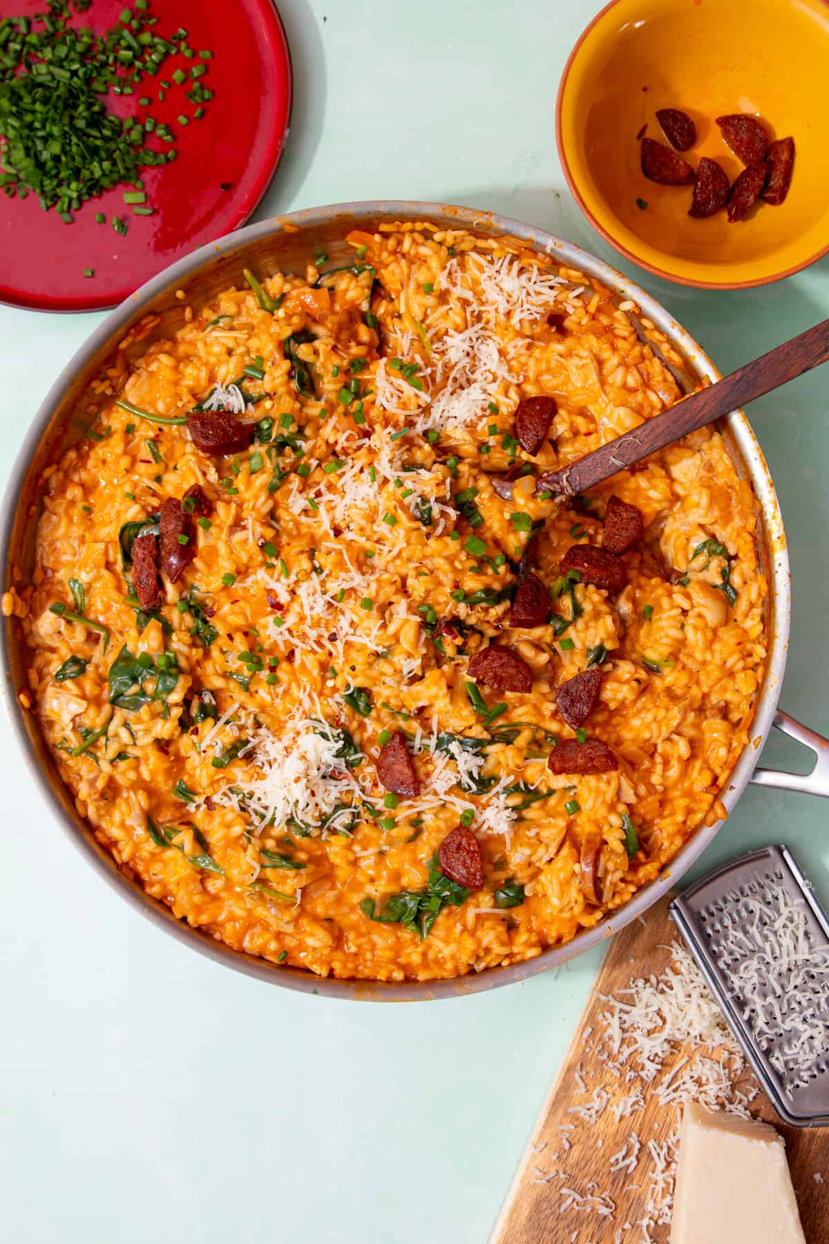 A large pan with risotto with chicken, topped pieces of chorizo,  chopped chives and parmesan with a wooden spoon. Small plates with chives, chorizo and grated parmesan on a board are next to pan.