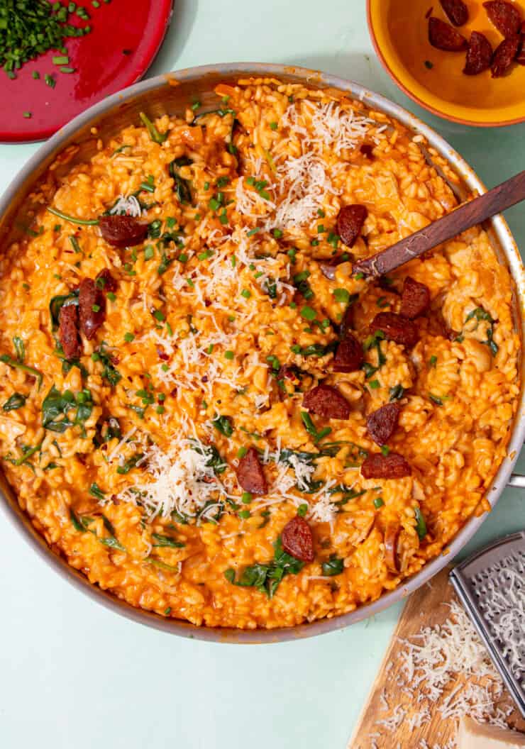 A large pan with choicken and chorizo risotto topped with parmesan, chorizo pieces and chives with a wooden spoon and small dishes of chives and chorizo next to the pan.