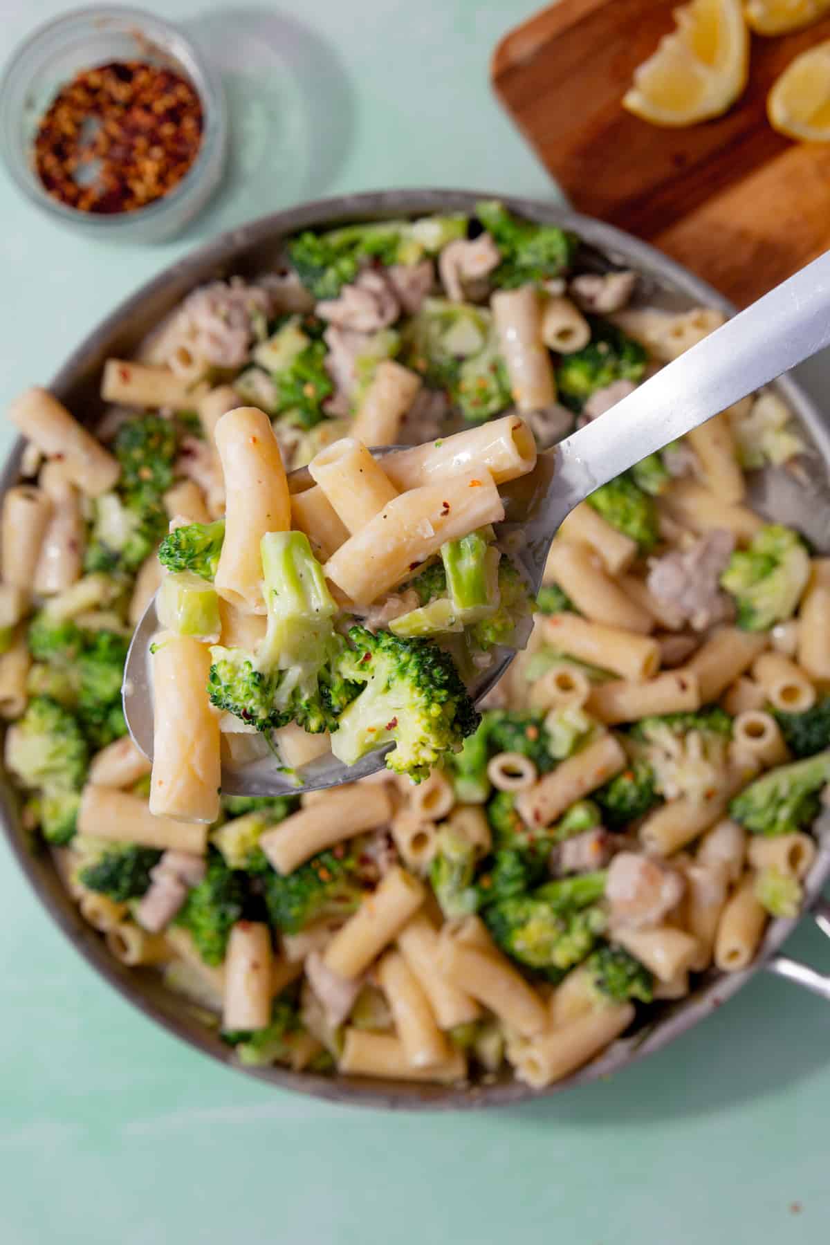 A large pan with broccoli, rigatoni pasta and chicken pieces, with a large metal spoon of the pasta showing above the pan.  Lemon wedges and chilli flakes in partial veiw behind the pan.