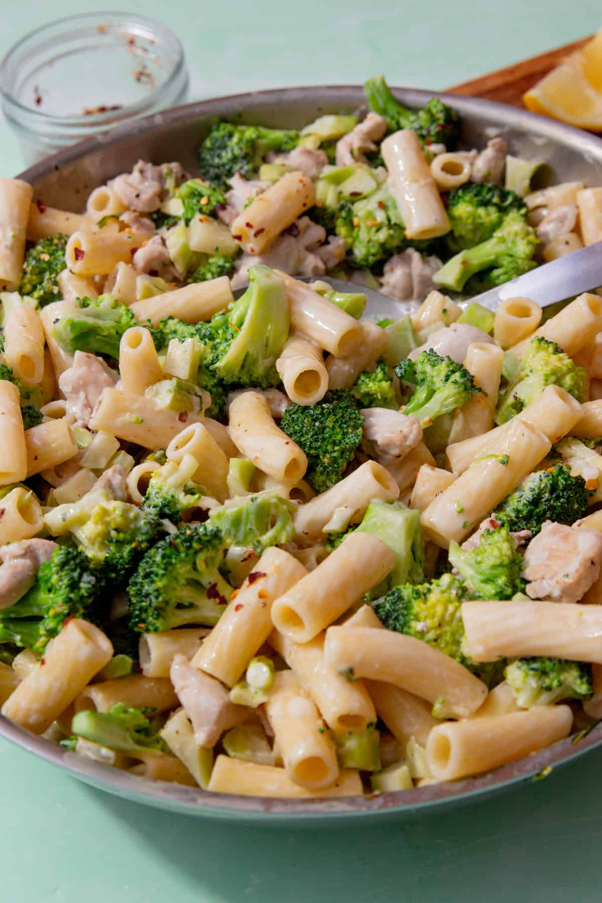 A large pan with broccoli, rigatoni pasta and chicken pieces, with a large metal spoon in the pasta.  