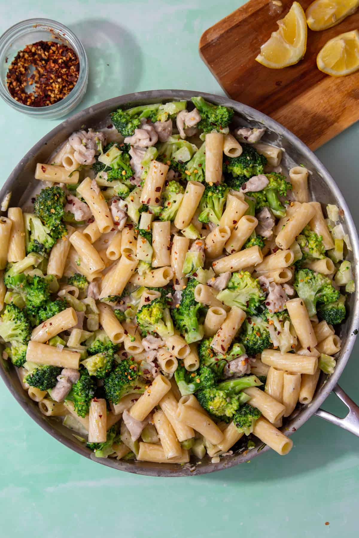A large pan with broccoli, rigatoni pasta and chicken pieces with lemon wedges on a chopping board and chilli flakes in a pot in partial veiw behind the pan.