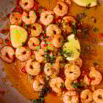 A large pan with King prawns in a sauce with lemon wedges, fresh parsley and red chillies.
