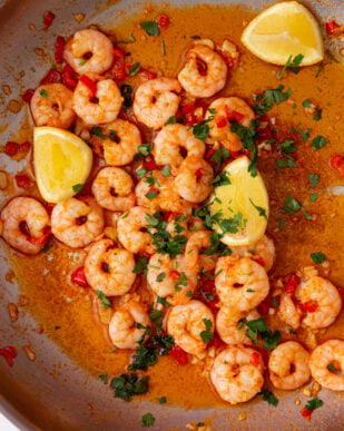 A large pan with King prawns in a sauce with lemon wedges, fresh parsley and red chillies.