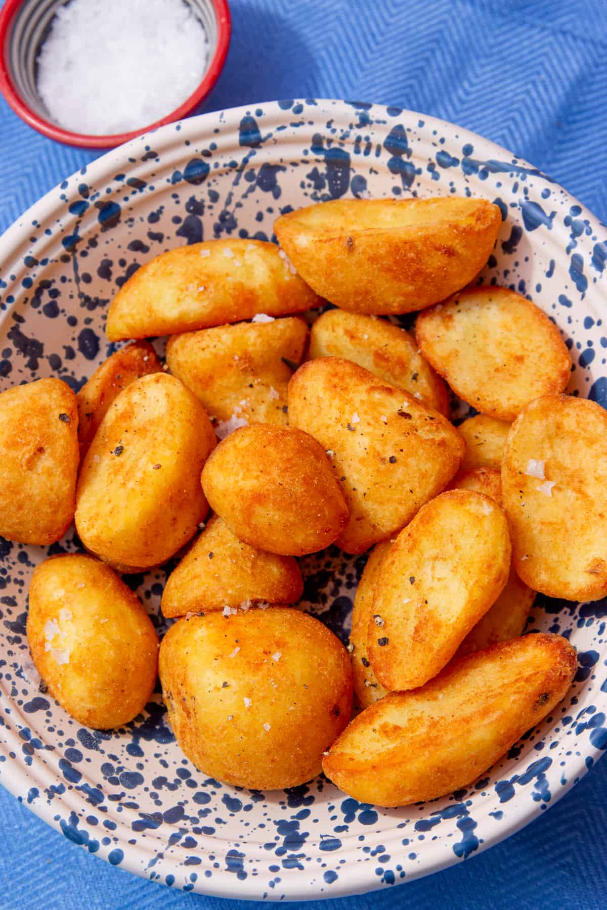 Golden browned roast potatoes in a patterned white and bowl with a small bowl of salt behind on a blue cloth.