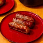 3 cooked and golden browned sausages on a red plate with another red plate in partial view.