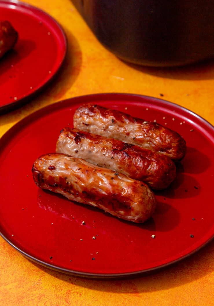 3 cooked and golden browned sausages on a red plate with another red plate in partial view.