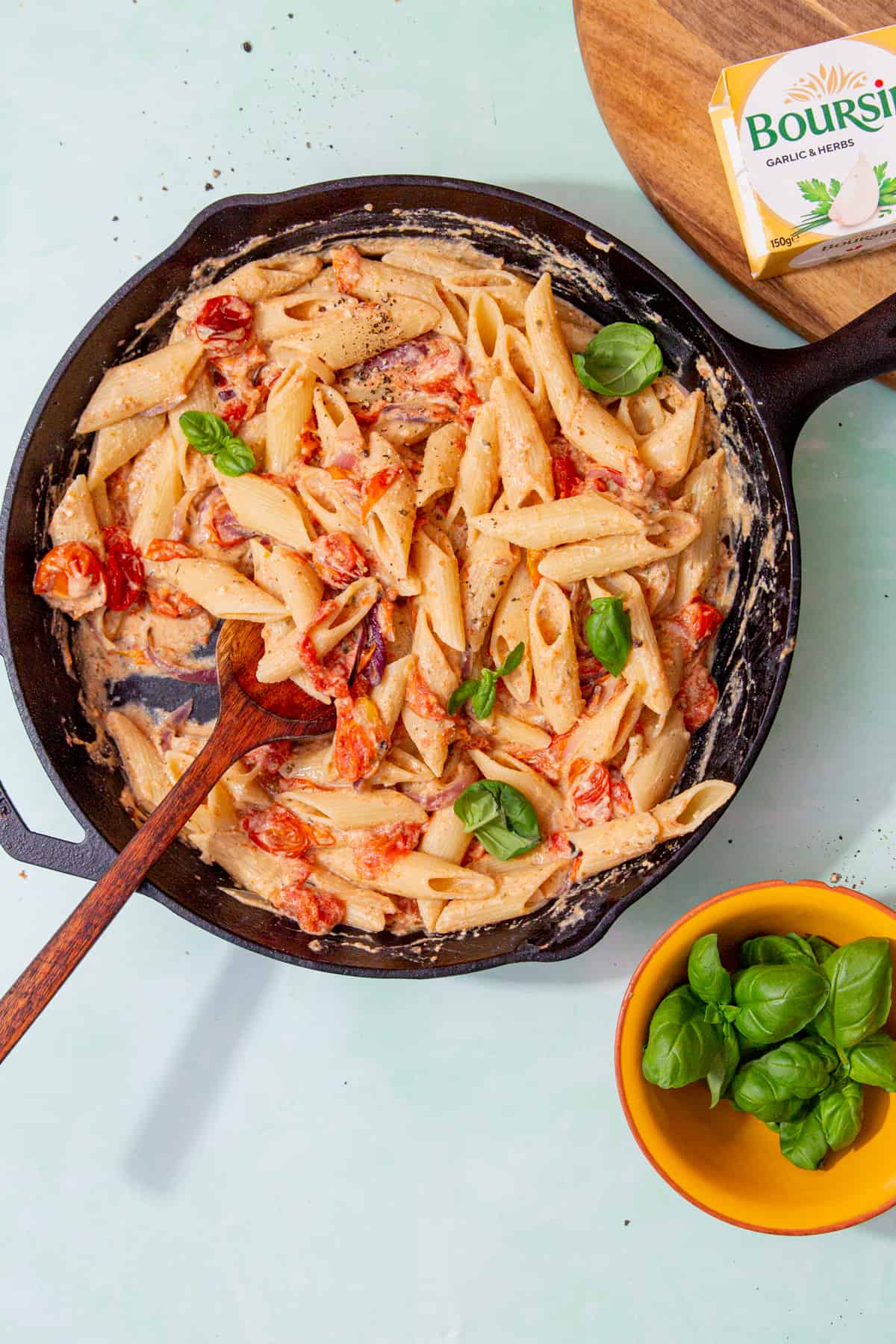 A large skillet pan with pasta with a creamy, cheesy sauce with cooked cherry tomatoes and a wooden spoon.  Boursin cheese package in view behind pan.