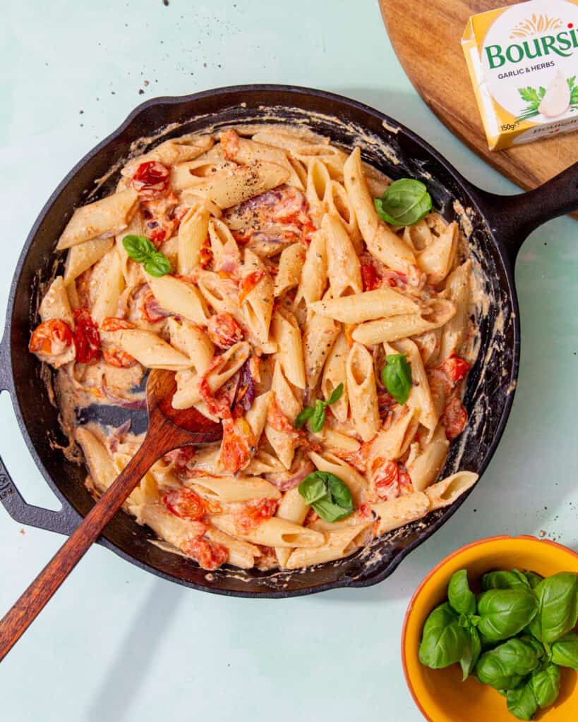 A large pan with pasta in a cheesy sauce, with cherry tomatoes and fresh basil next to a pack of Boursin cheese and fresh basil.