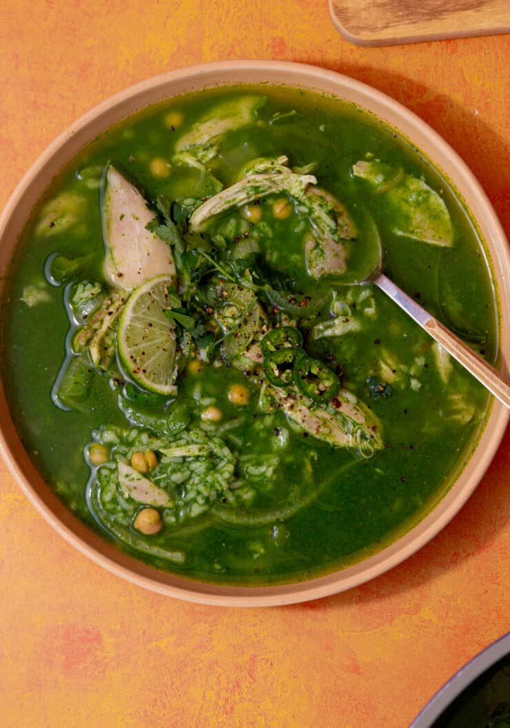 Green soup in a bowl, with chicken, rice and chillies and a wedge of lime.