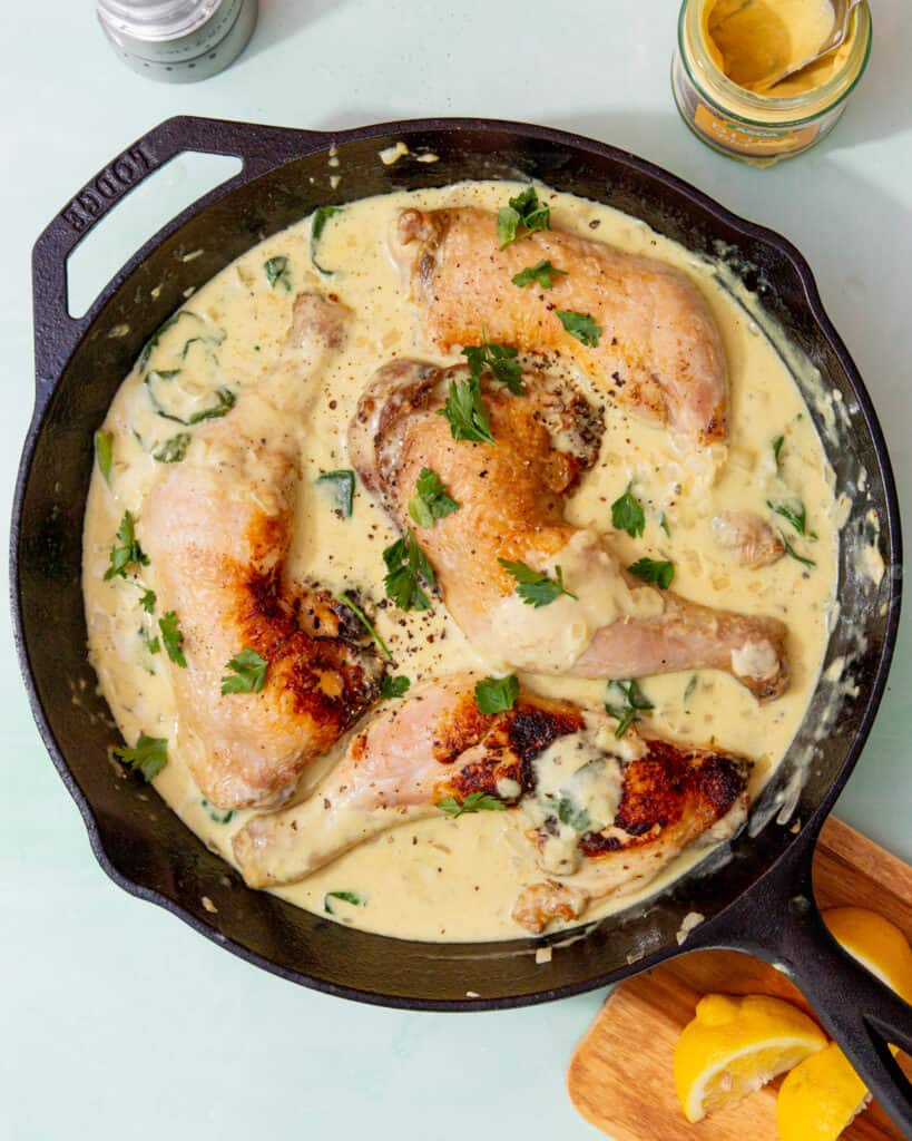 4 golden borwned chicken legs in a skillet pan covered with a creamy sauce and fresh herbs.