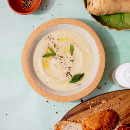 A creamy soup in a bowl with a little oil and fresh basil next to a loaf of bread, a parsnip and cream cheese also in partial view.