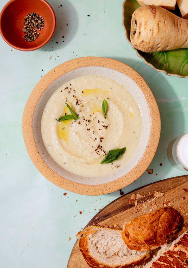 A creamy soup in a bowl with a little oil and fresh basil next to a loaf of bread, a parsnip and cream cheese also in partial view.