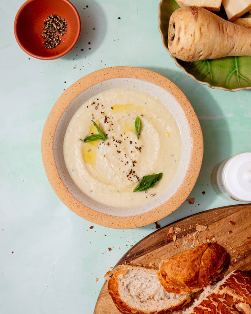 A creamy soup in a bowl with a little oil and fresh basil next to a loaf of bread, a parsnip and cream cheese also in partial view.