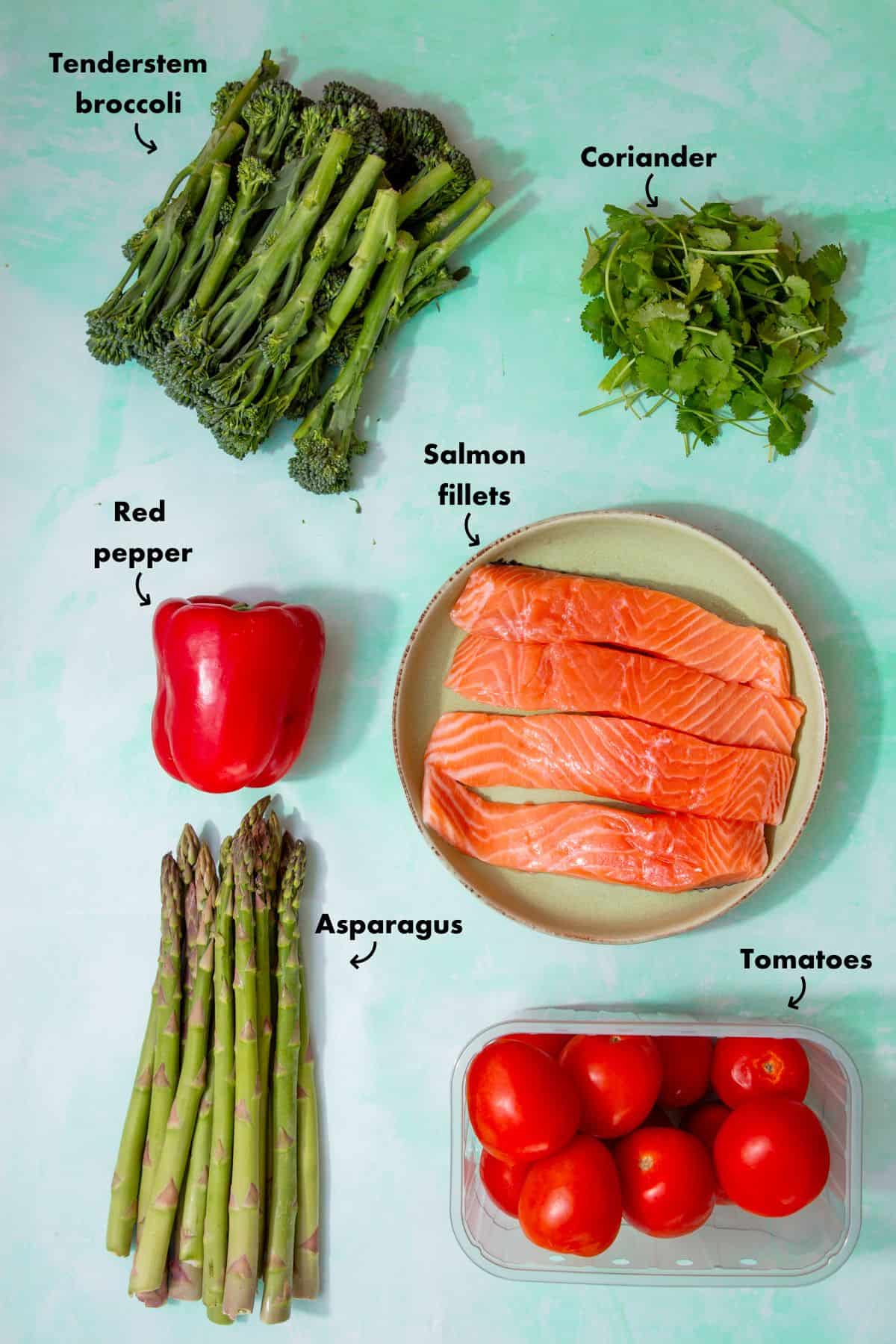 Ingredients to make a roasted salmon tray bake laid out on a plae blue background and labelled.