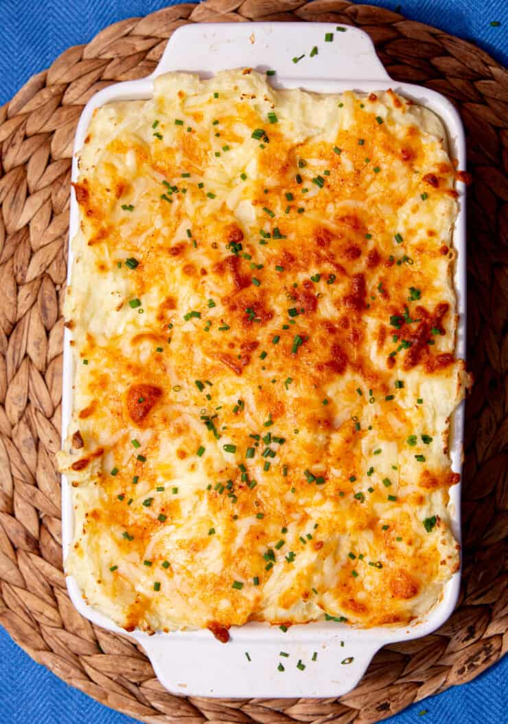 A rectangular white baking dish with mashed potatoes covered in ca golden browned cheesy topping garnished with fresh chives.