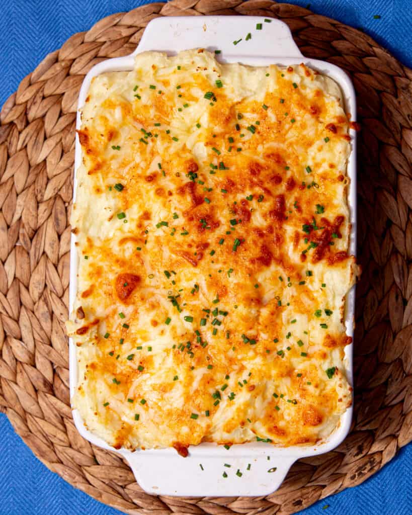 A rectangular white baking dish with mashed potatoes covered in ca golden browned cheesy topping garnished with fresh chives.