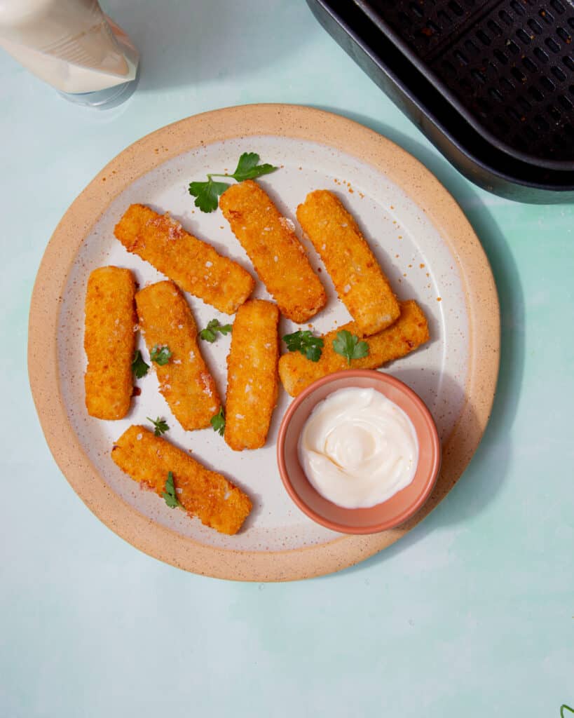 8 golden browned battered fish fingers on a plate with some mayonnaise in a small bowl and some fresh parsley.