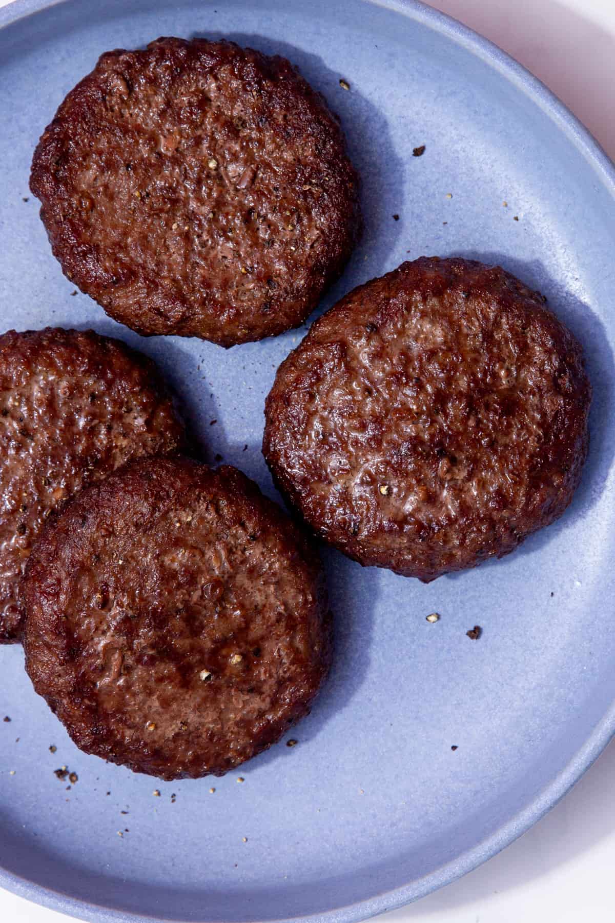 4 cooked, dark browned burgers on a blue plate with some pepper seasoning.