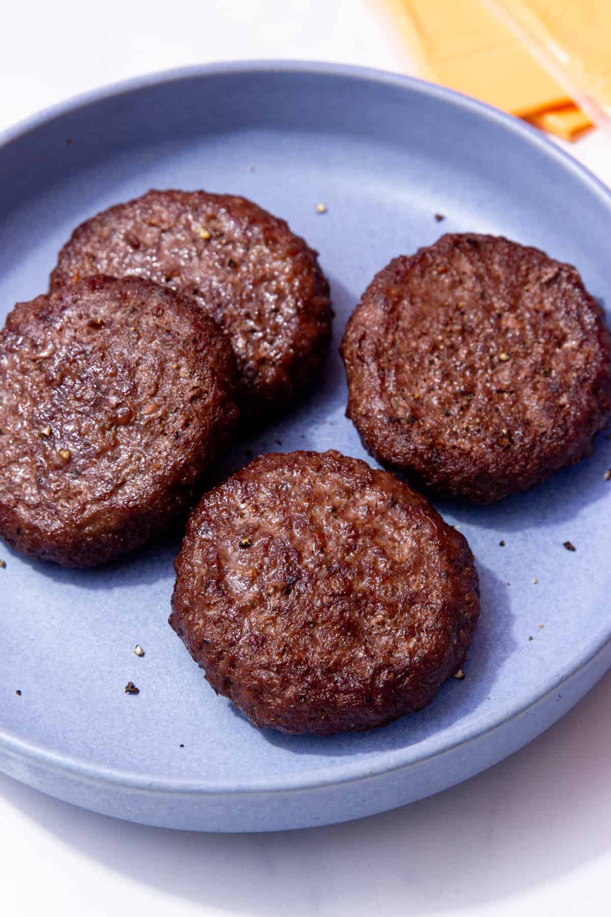 4 cooked, dark browned burgers on a blue plate with some yellow cheese slices in partial view.