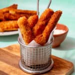 A metal container with halloumi fries in parchment paper and more fries on a plate behind.