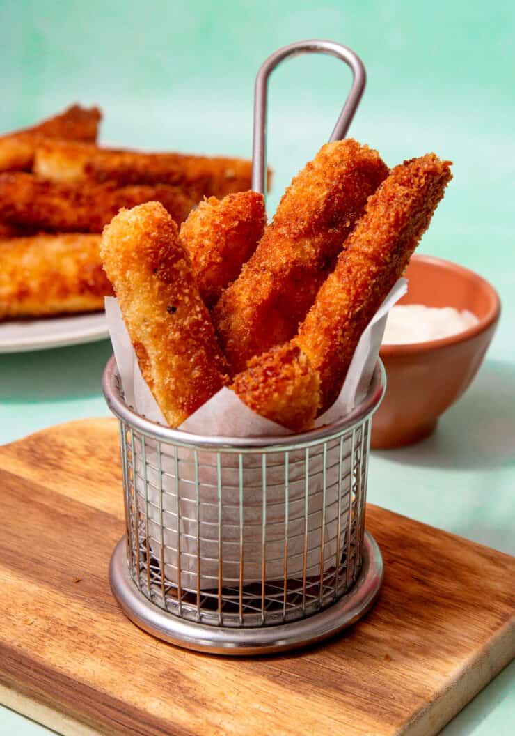 A metal container with halloumi fries in parchment paper and more fries on a plate behind.