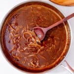 A large pan with gravy and onions with a wooden spoon and some sprigs of rosemary in partial view.