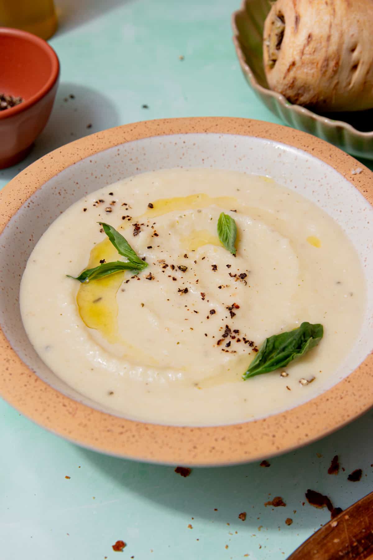 A bowl of creamy pale coloured soup topped with fresh basil and some oli and seasoning with a parsnip in partial view behind.