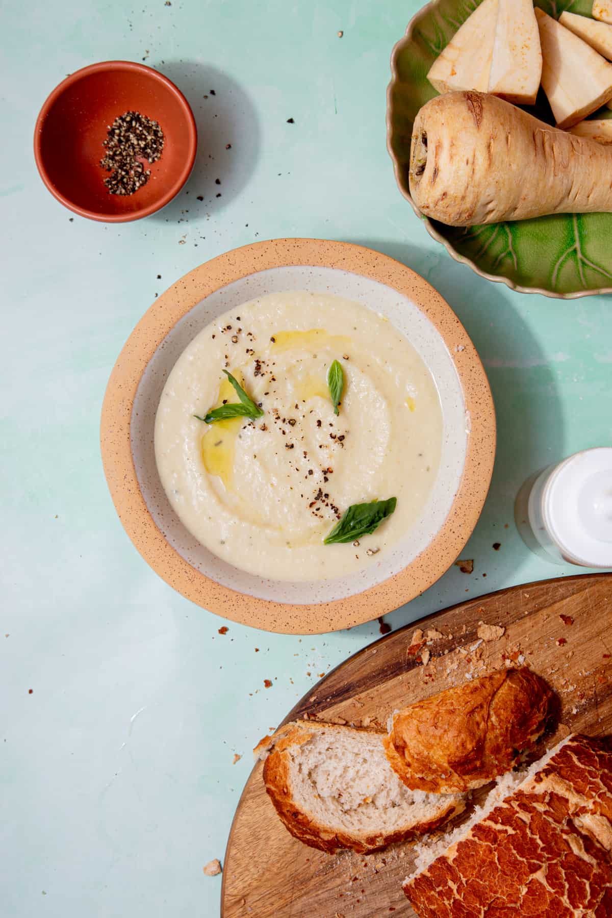 A bowl of creamy pale coloured soup topped with fresh basil and some oli and seasoning with a crusty loaf sliced on a chopping board and some parsnips on a plate.