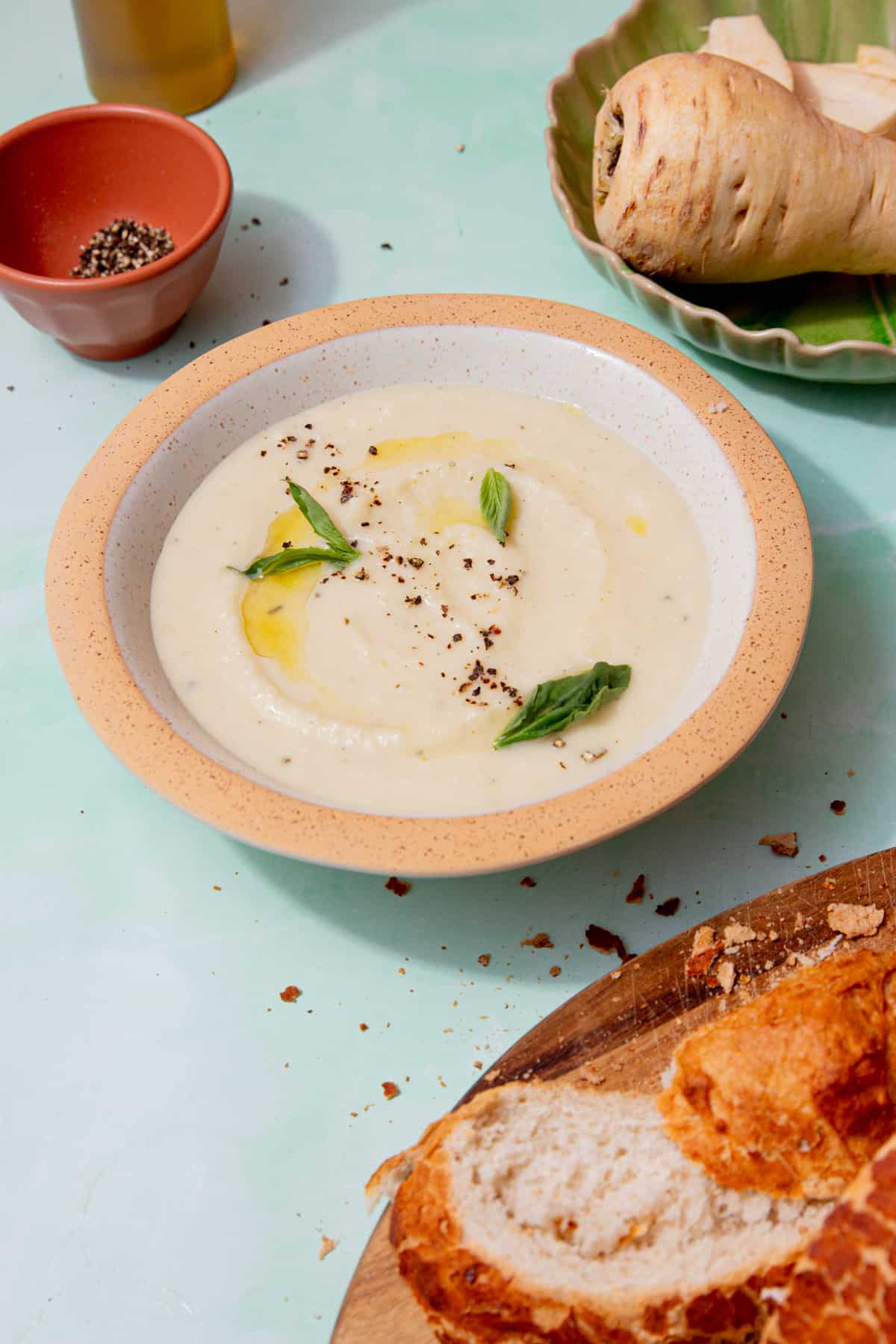 A bowl of creamy pale coloured soup topped with fresh basil and some oil and seasoning with a crusty loaf sliced on a board and some parsnips on a plate behind.