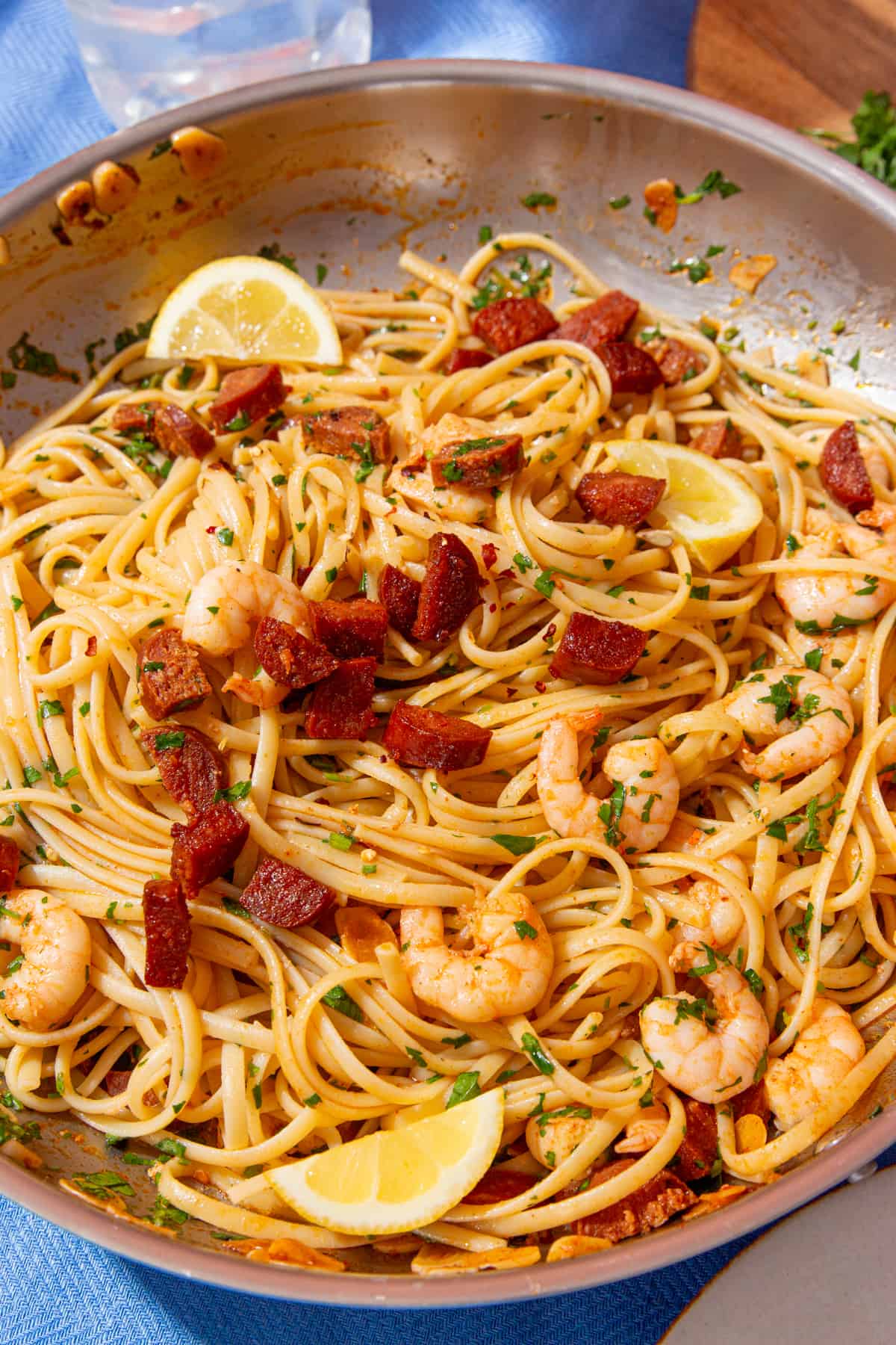 Close up of king prawns and chorizo in linguine with green herbs and lemon wedges in a large pan.