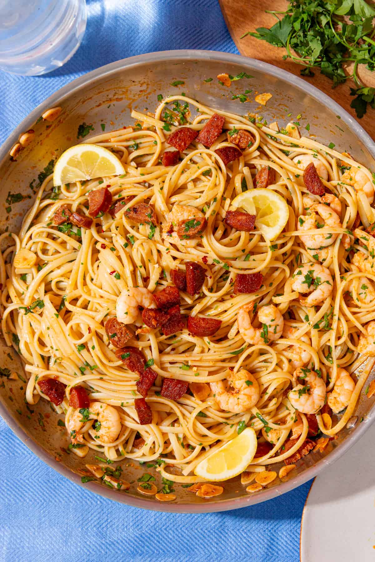 Close up of king prawns and chorizo in linguine with green herbs and lemon wedges in a large pan on a blue background with some parsley in partial view.
