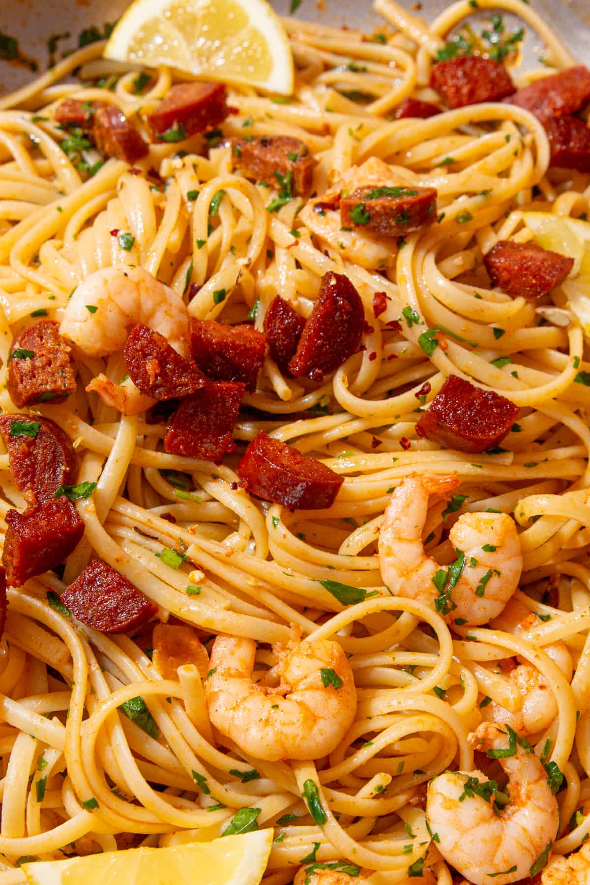 Close up of king prawns and chorizo in linguine with green herbs and a lemon wedge.