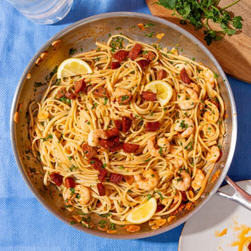 A large stainless steel pan with linguine, chorizo and king prawns with green herbs and topped with lemon wedges on a blue background.