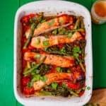 A white tray with baked salmon, and asparagus with other vegetables on a green background.