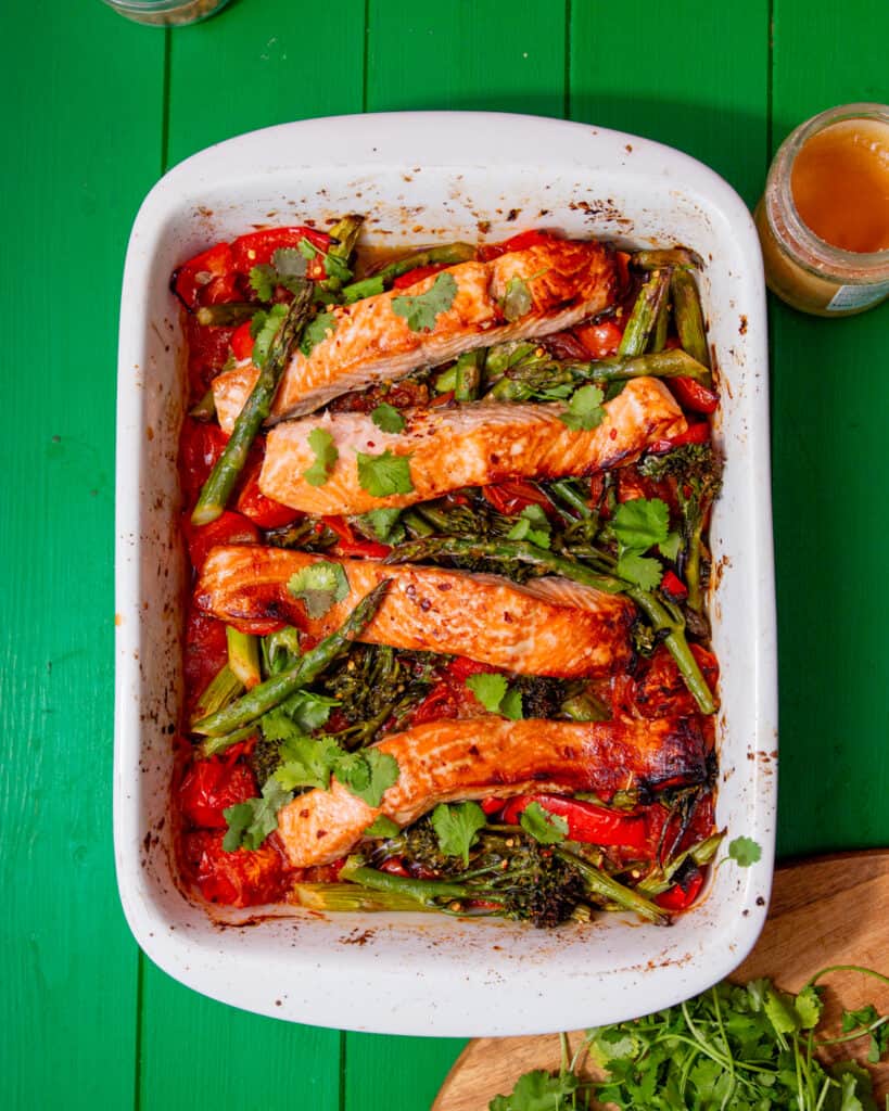 A white tray with baked salmon, and asparagus with other vegetables on a green background.