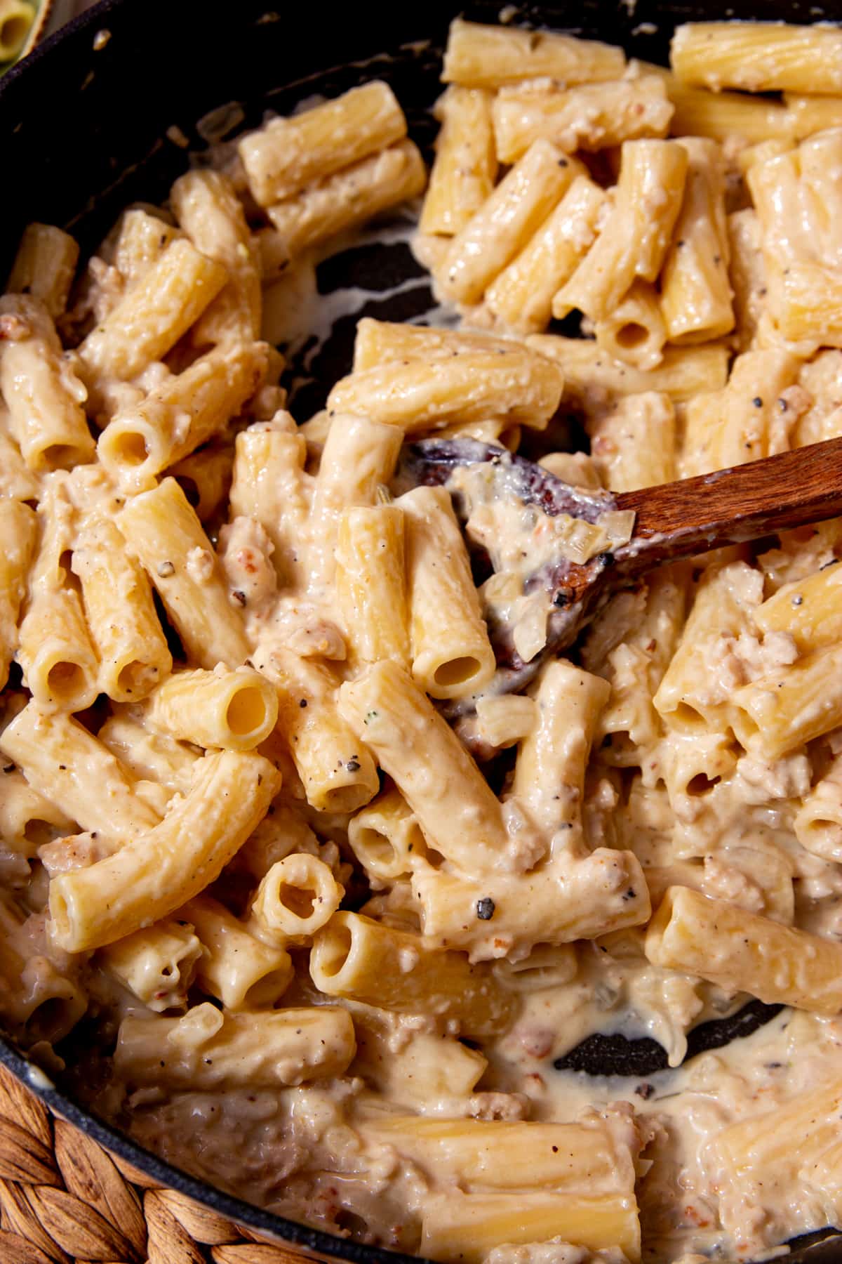 Creamy rigatoni cooked pasta with sausage and bits in a pan being stirred with a wooden spoon.
