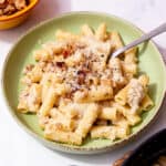 A bowl of rigatoni pasta in a creamy sauce topped with grated parmesan and sausage bits with a spoon.