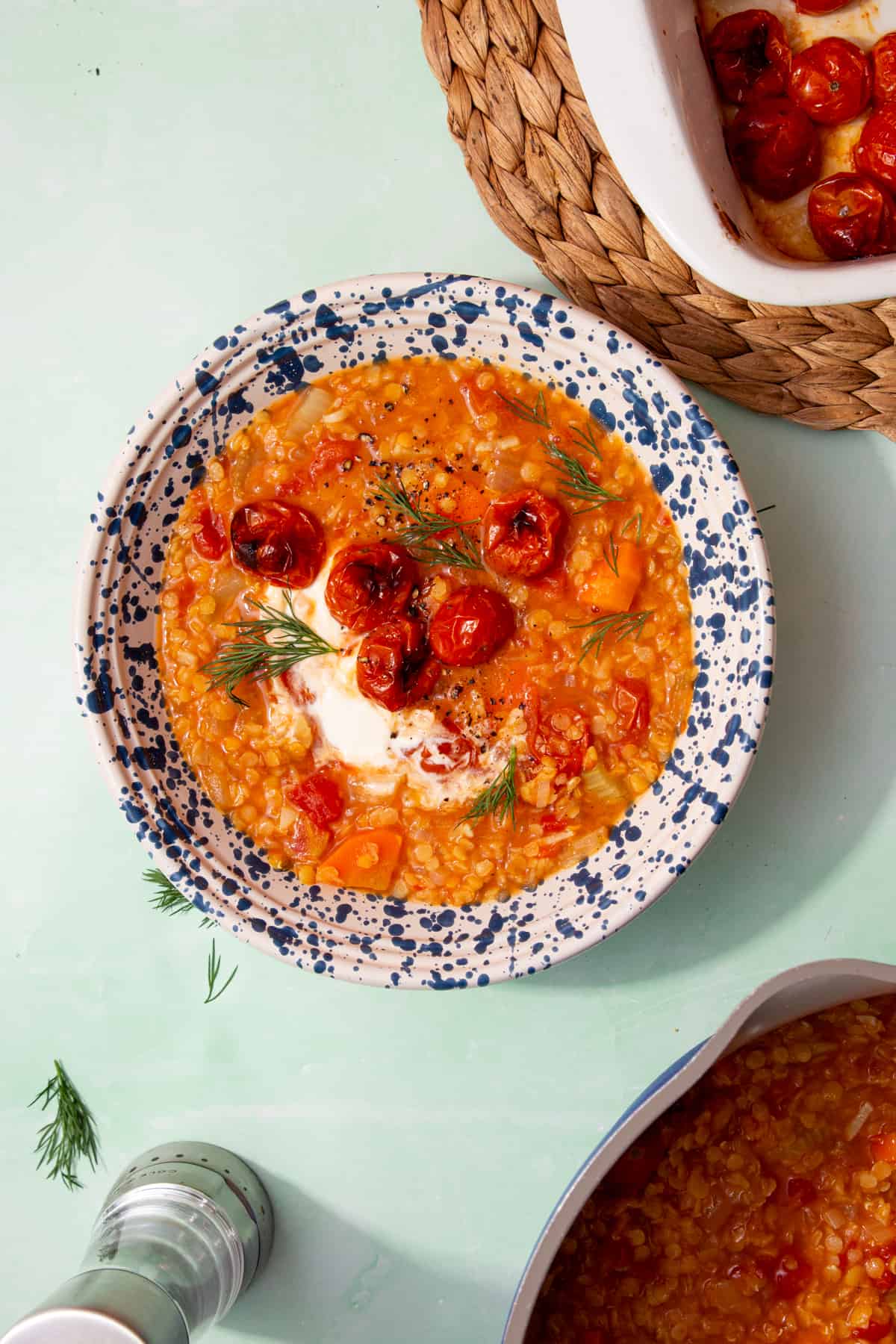 A patterned bowl full of red lentil soup with roasted cherry tomatoes, some dill and a dollop of creme fraiche with a baking tray with roasted tomatoes in partial view.