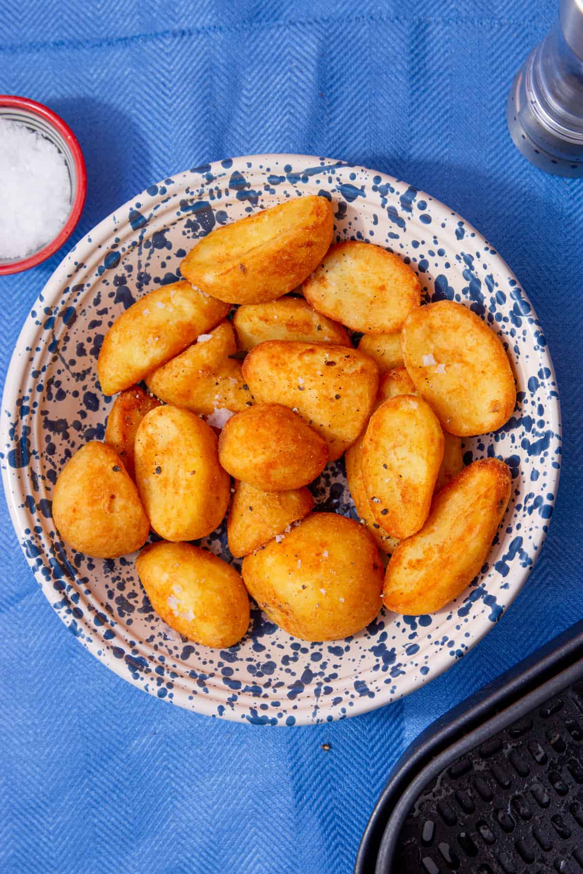 Seasoned golden browned roast potatoes in a patterned white and bowl with a small bowl of salt behind on a blue cloth.
