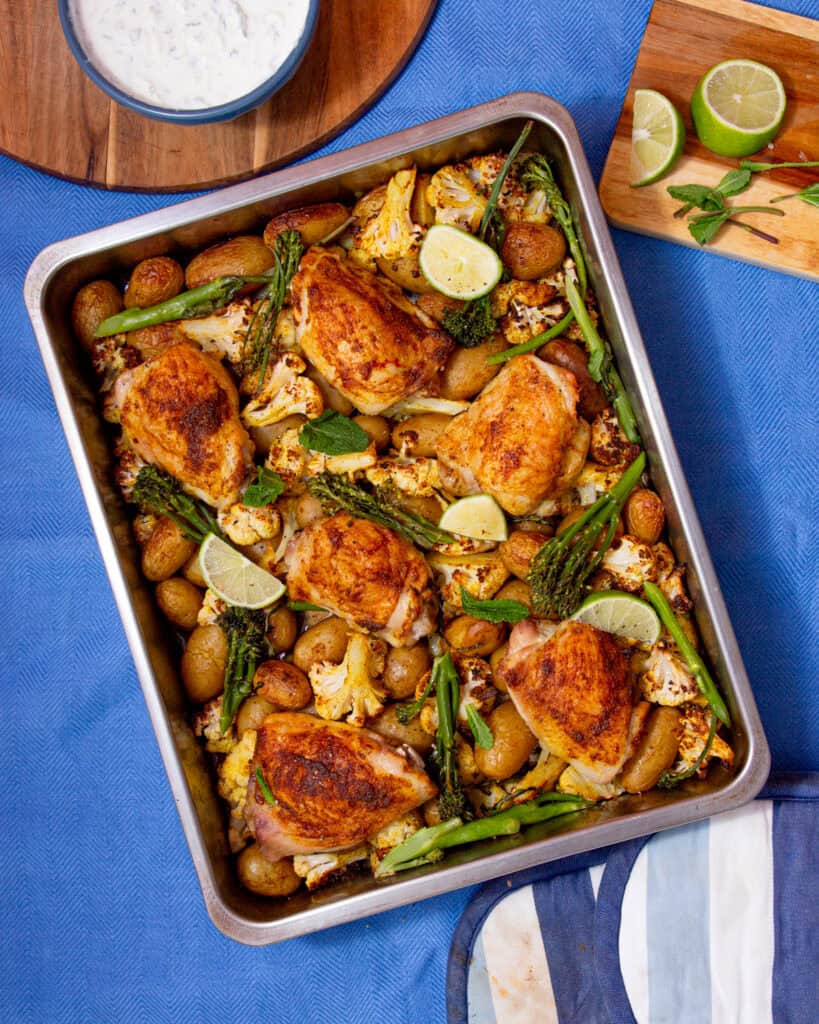 A baking tray with golden browned chicken thighs, cauliflower and broccoli with some lime wedges next to a bowl of raita and lime wedges.