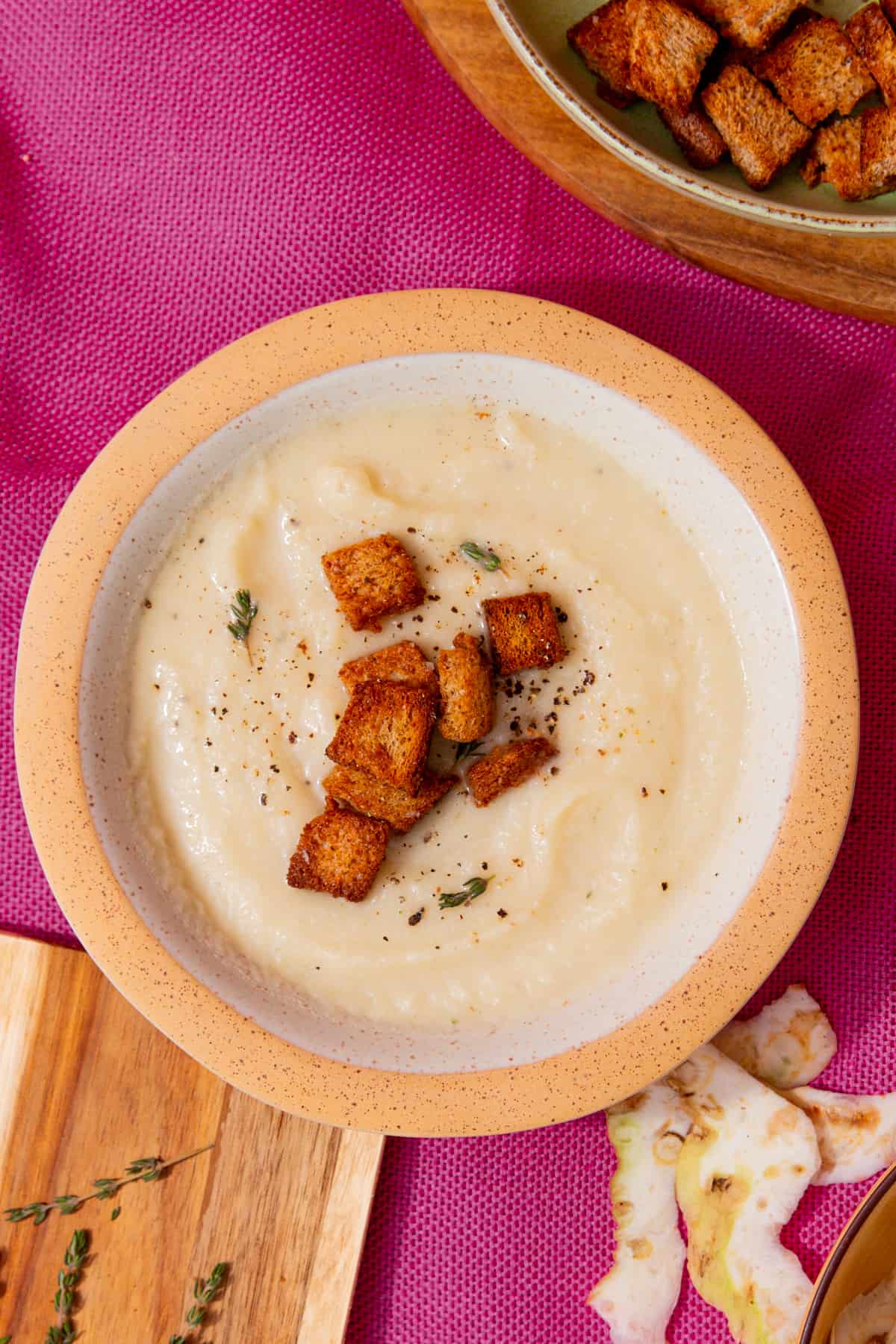 A bowl of pale creamy soup with golden browned croutons and seasoning in a bowl on a pink background next to some extra croutons in partial view, celeriac peel on and thyme sprigs.