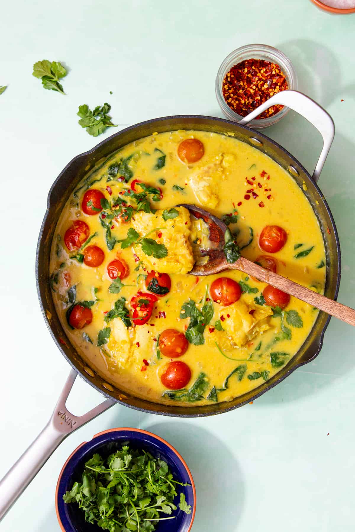 A large pan with a yellowish curry with fish on a wooden spoon showing cherry tomatoes and fresh coriander next to a small bowl of chilli flakes and another small bowl with fresh coriander.
