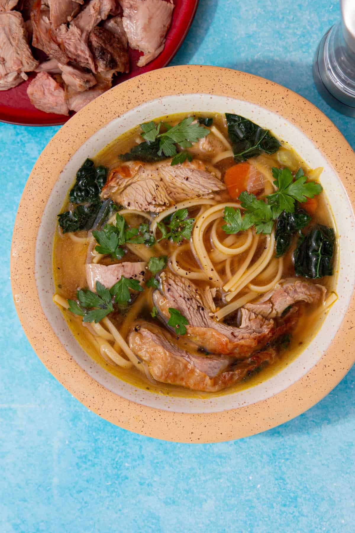 A bowl of soup with noodles, turkey, cavolo nero, carrot and some parsley with the turkey on a plate in partial view on a blue background.