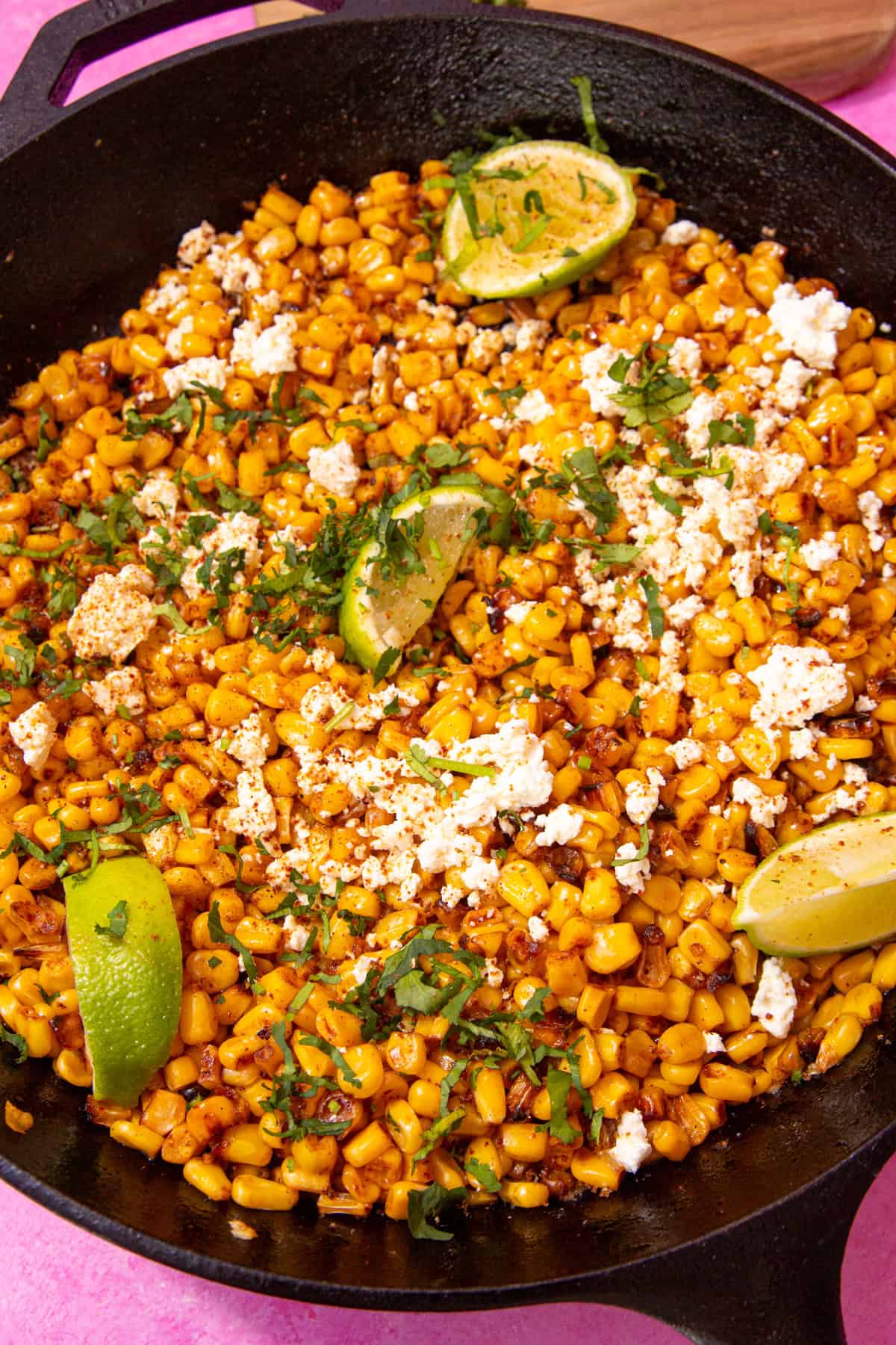 A pan with golden browned sweet corn, topped with crumbled feta, lime wedges and fresh chopped coriander.