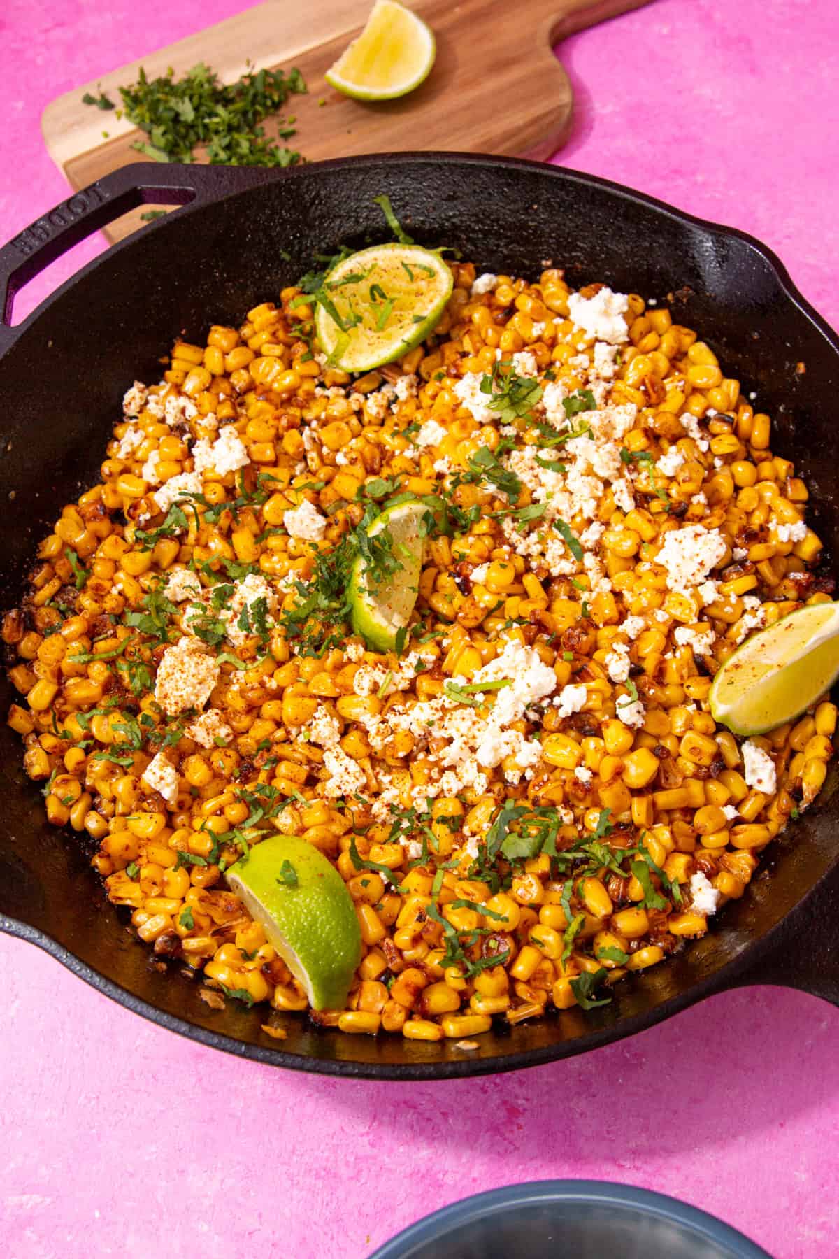 A pan with golden browned sweet corn, topped with crumbled feta, lime wedges and fresh chopped coriander with a small chopping board with toppings behind.